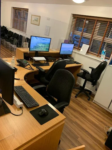 Modern office with wooden desks and black chairs, featuring multiple computer monitors. Warm lighting and blinds enhance the inviting atmosphere.